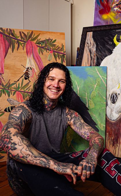 Photo of Indigenous artist Josh Deane sitting in front of his paintings in a studio